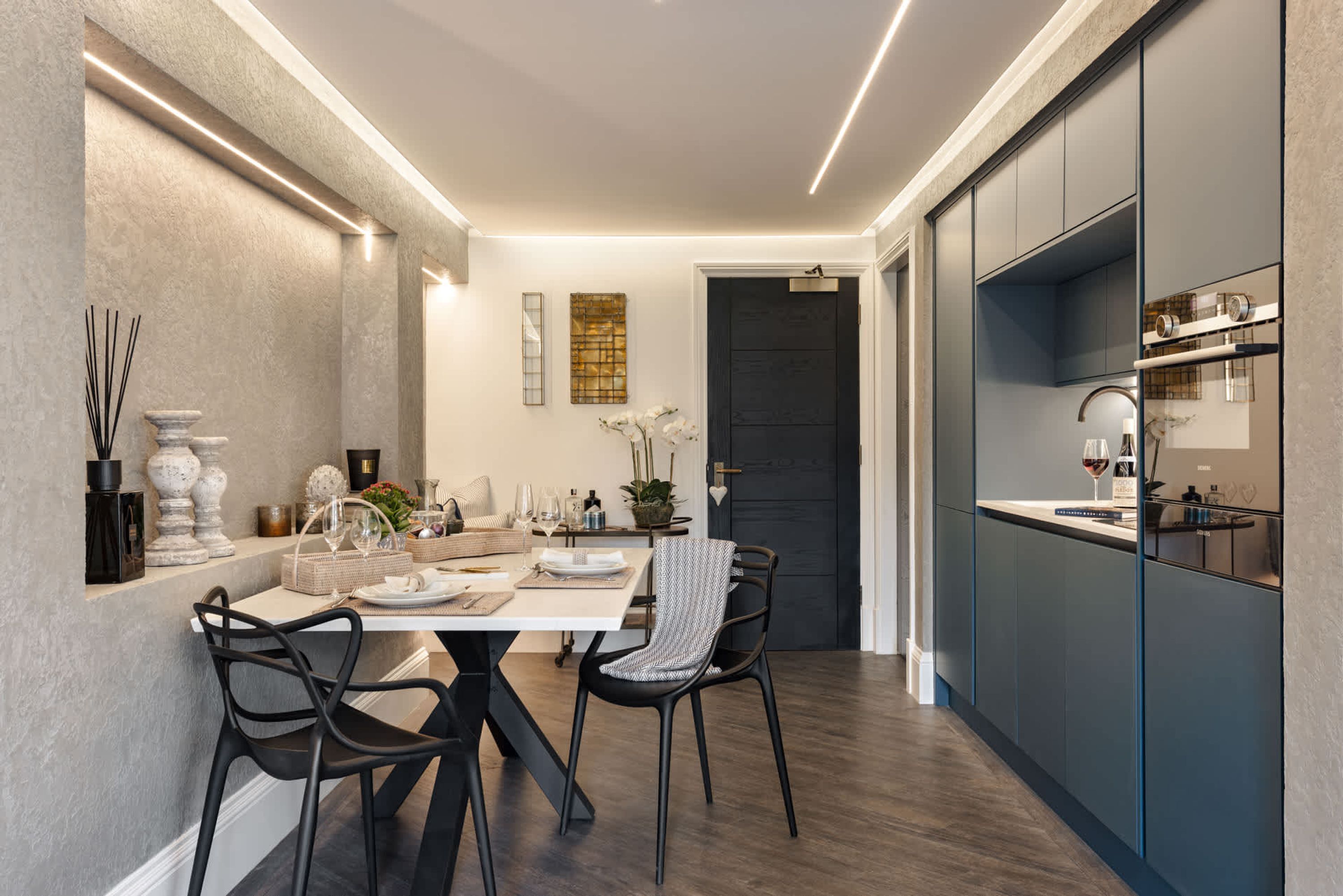 A modern kitchen with dark cabinetry, a dining area with a white table and black chairs, and a dark door in the background.