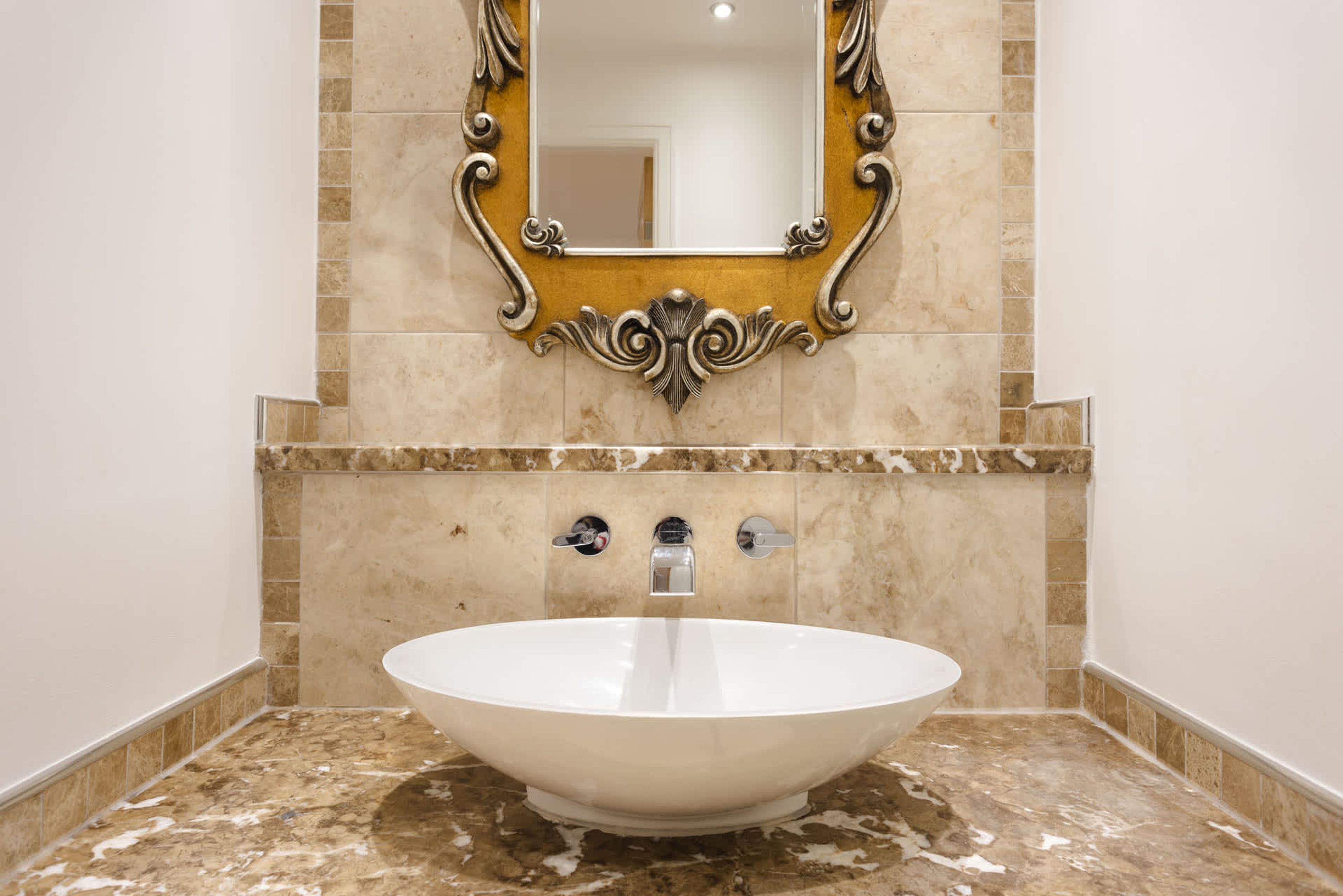 A bathroom with a large oval sink, a mirror with ornate gold detailing, and a tiled wall.