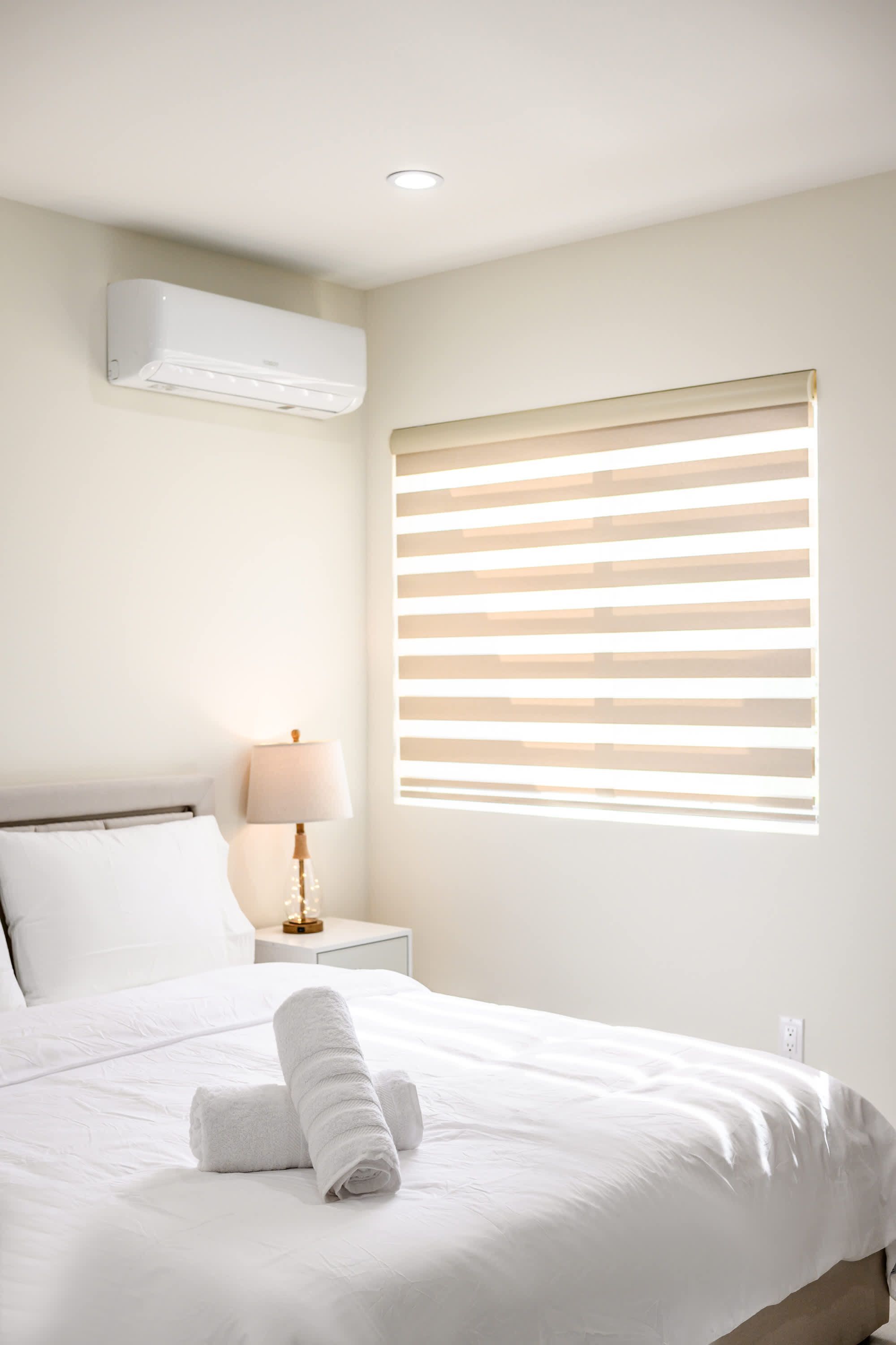A neatly made bed with white linens and a rolled towel, a bedside lamp, and a window with horizontal blinds.