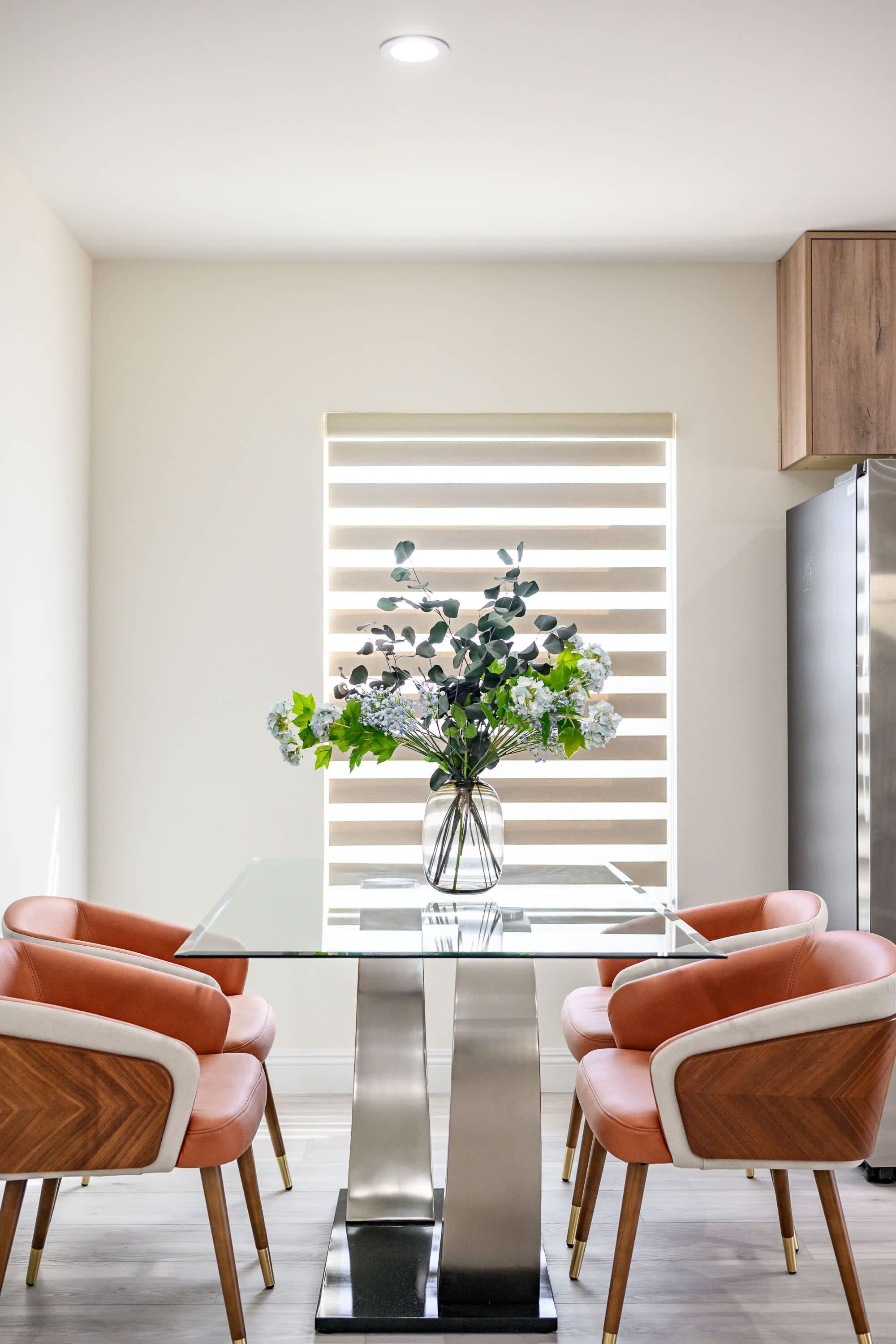 A modern kitchen with a glass dining table, two orange chairs, a stainless steel refrigerator, and a vase with flowers on the table.