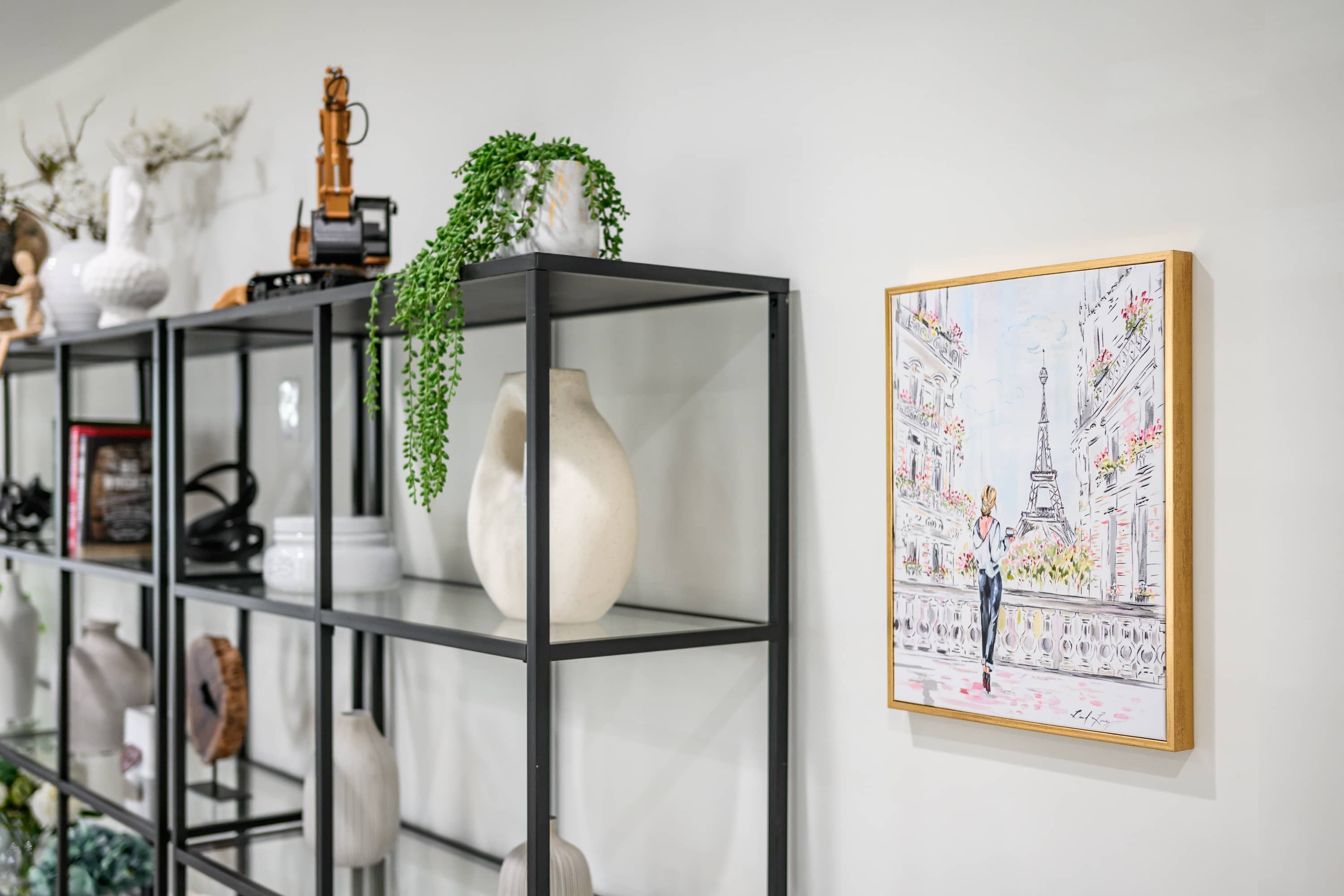 A modern interior with a black metal shelving unit displaying various decorative items and a framed illustration of a Parisian street scene.