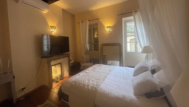 A bedroom with a bed, a television on a stand, a fireplace, and a window with curtains. - Magnificent apartment with direct access to the Place des 9 Jets in CERET. (Céret)