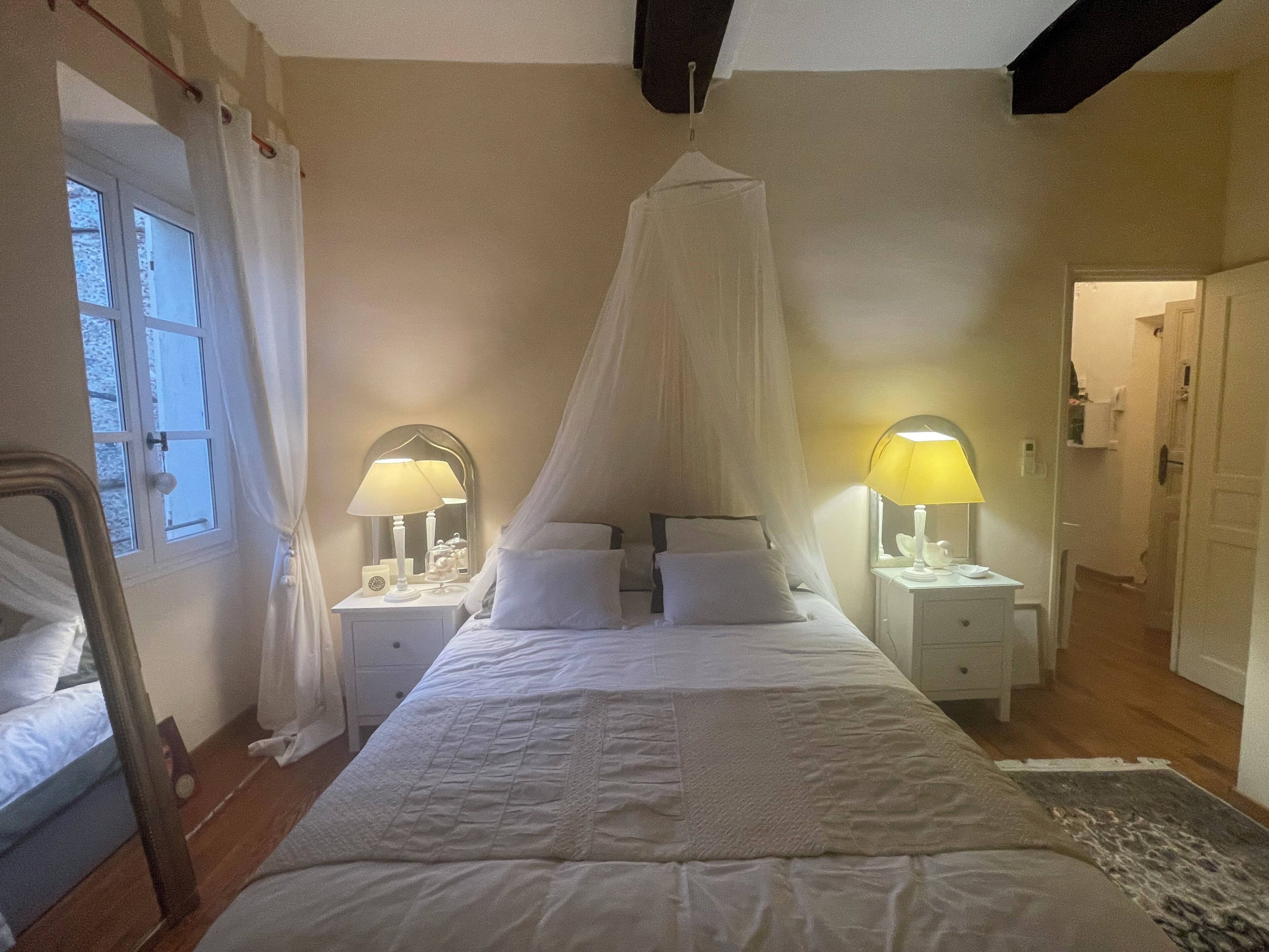 A bedroom with a large bed, two bedside tables with lamps, a window with curtains, and a door leading to another room.