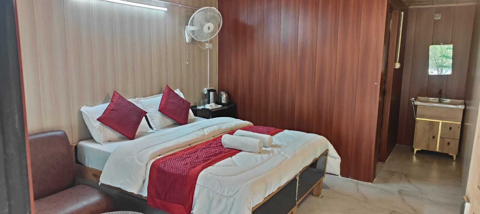 A bedroom with a bed, pillows, a bedside table with a fan, a nightstand, and a view of a bathroom.