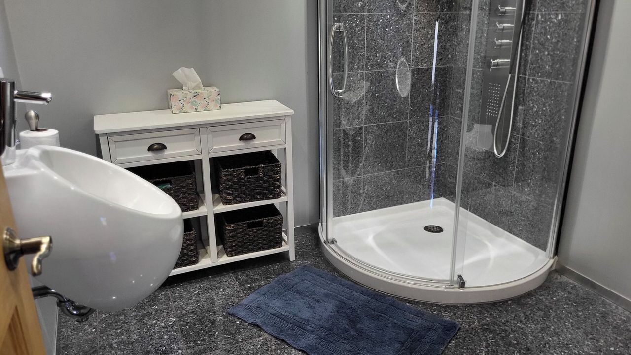 Un baño con un lavabo blanco, un tocador blanco con cajones, un box de ducha de vidrio y una alfombra azul en el suelo.