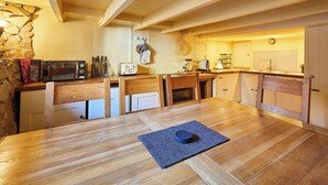 Una mesa de comedor de madera con un mantel individual y un tazón, rodeada de sillas de madera. Se observan en el fondo una pared de piedra y vigas de madera en el techo.