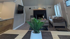 Amplia sala de estar con techo alto, vigas de madera, chimenea, televisor y mesa de comedor con una planta.