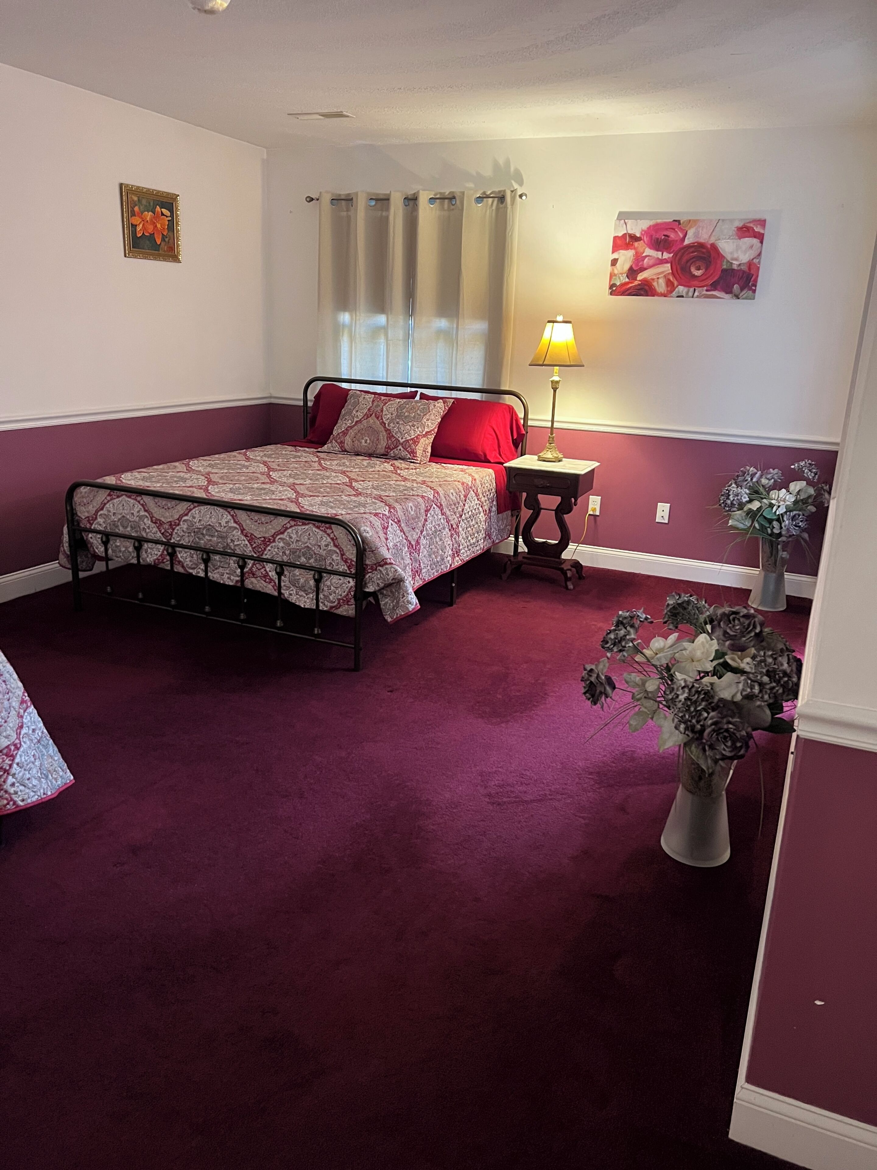 A bedroom with a bed, a nightstand, a lamp, a vase with flowers, and a painting on the wall.