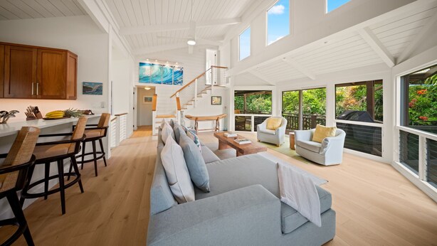 A spacious living area with a large gray sofa, wooden dining table, and chairs. A staircase leads to an upper level with large windows. - Hale Kailea, Beautiful Hanalei Home TVNC#4241 (Hanalei)