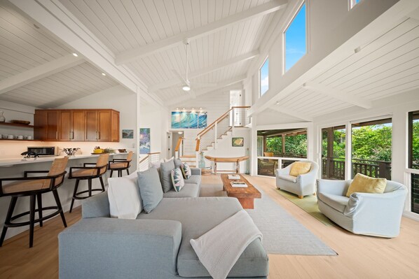 A spacious living area with a large gray sofa, a wooden coffee table, and a white armchair. There is a kitchen area with wooden cabinets and a bar stool. A staircase leads to an upper level with large windows.
