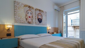 Een hotelkamer met een bed, twee wandlampen, een raam met uitzicht op een balkon en twee schilderijen aan de muur.