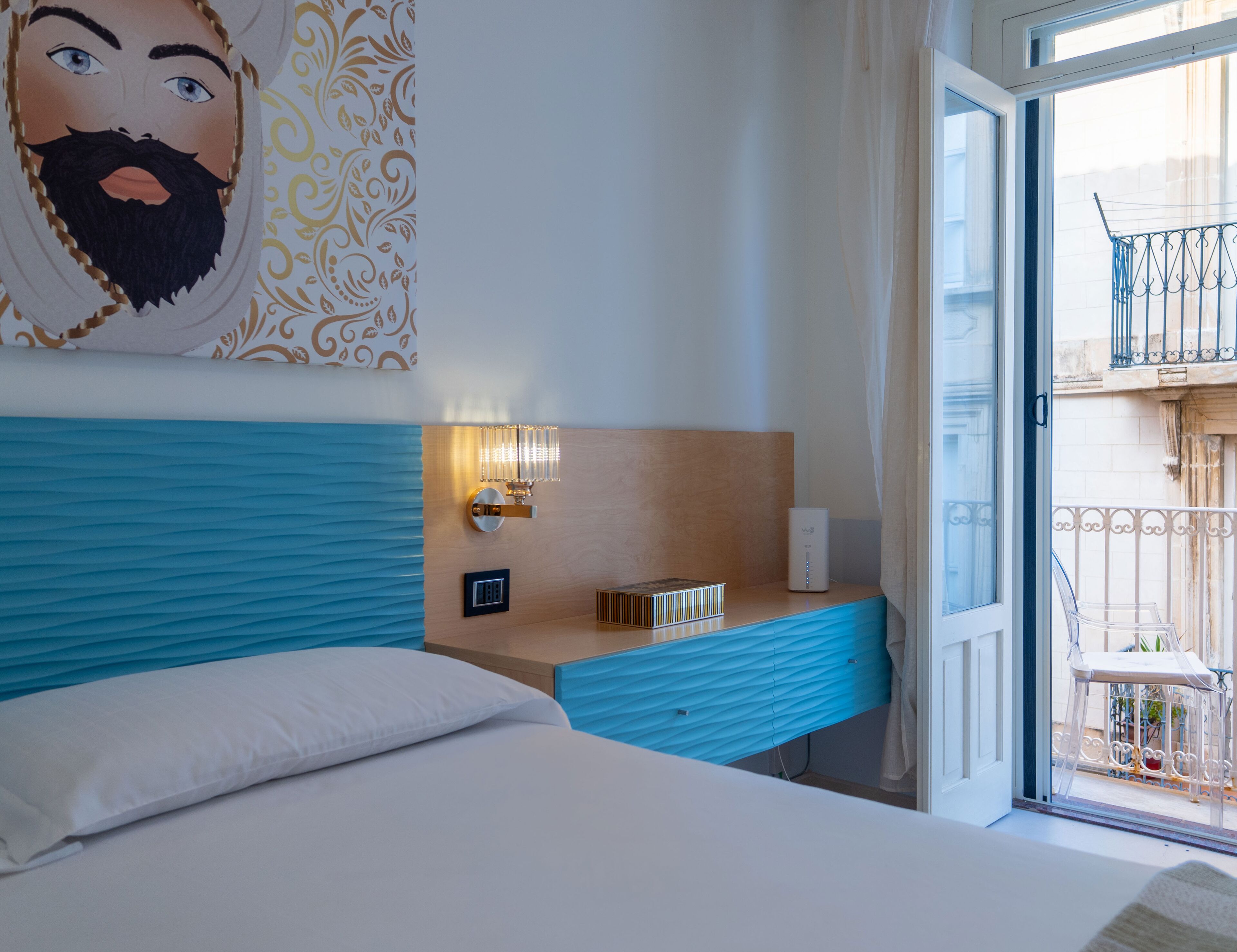A hotel room with a bed, a bedside table, a wall-mounted light, a power outlet, and a view of the outside balcony.