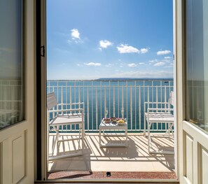En balkon med to hvide stole, et lille bord med mad og drikke, og havudsigt.