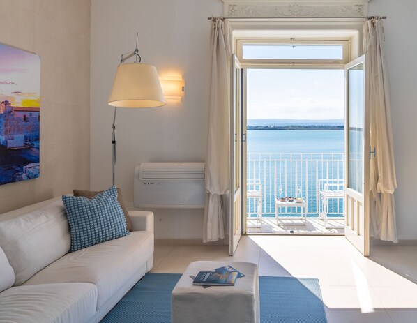 Et værelse med en hvid sofa, pude med blåt mønster, gulvlampe, et lille bord med en bog og en balkon med havudsigt.