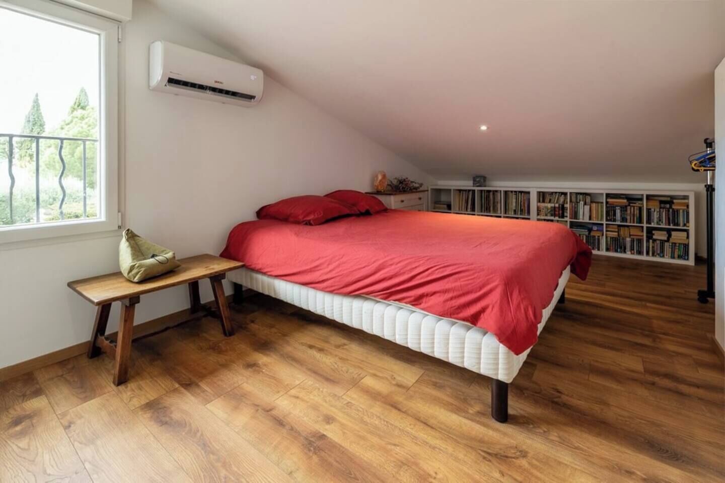 Ein Schlafzimmer mit Bett, einer Bank, einer Klimaanlage, einem Fenster und einem Bücherregal.