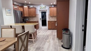 A modern kitchen with wooden cabinets, a stainless steel refrigerator, a dining area with a wooden table and chairs, and a trash can.