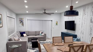 A modern living room with a grey sectional sofa, a glass coffee table, a wooden dining table with chairs, a ceiling fan, and a mounted TV.