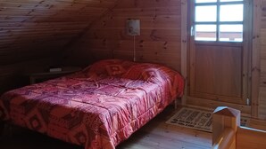 Una habitación pequeña y acogedora con techo inclinado, una cama individual con manta estampada, una puerta de madera con ventana y una mesita.
