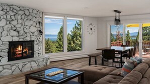A living room with a stone fireplace, large windows, a dining area, and a view of the outdoors. - Hch1083 - Mountain Serenity 4 Bedroom Home (South Lake Tahoe)