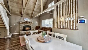A spacious living room with a fireplace, wooden ceiling beams, a dining table with chairs, and a large window. - Cyh0876 - Island Cabin 3 Bedroom Cabin (South Lake Tahoe)