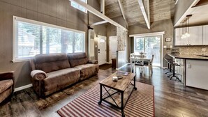 A spacious living area with a large brown leather sofa, a wooden coffee table, and a dining table with chairs. The room has high ceilings with exposed beams and large windows. - Cyh0876 - Island Cabin 3 Bedroom Cabin (South Lake Tahoe)