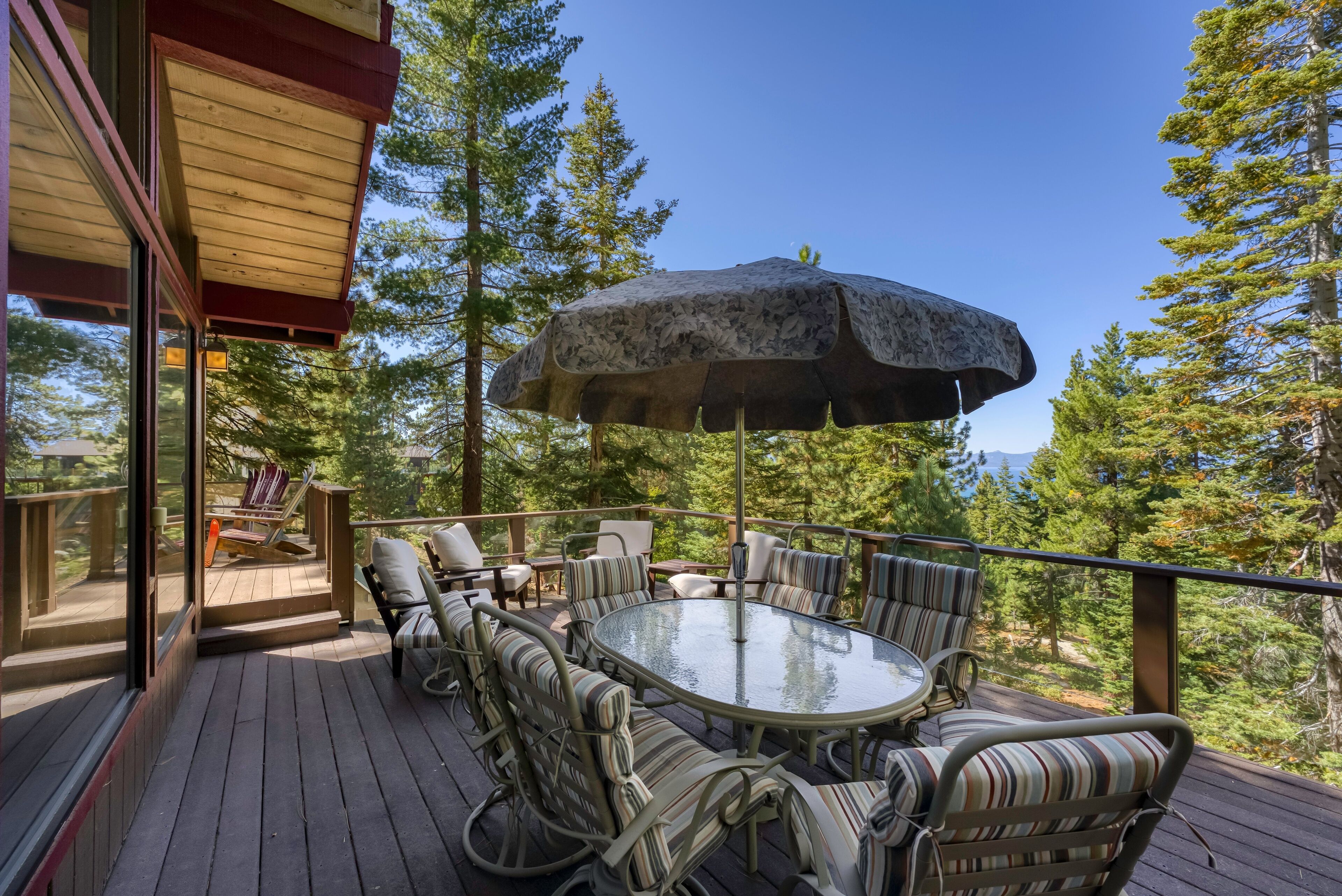 A deck with a glass-top table and striped chairs, surrounded by tall trees and a clear sky.