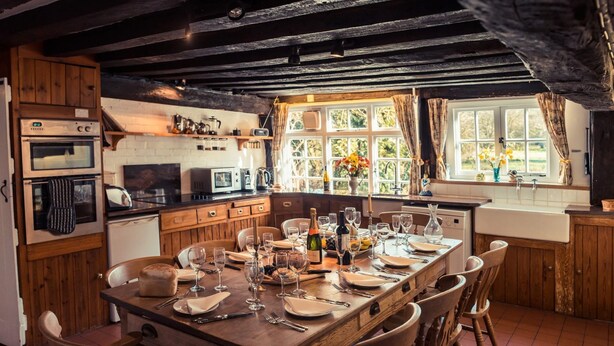 A rustic kitchen with wooden cabinets, a dining table set for six, and a large window with curtains. - Toad Hall Swilland, a beautiful, 5 bedroom country residence surrounded by Suffolk countryside (Swilland,)