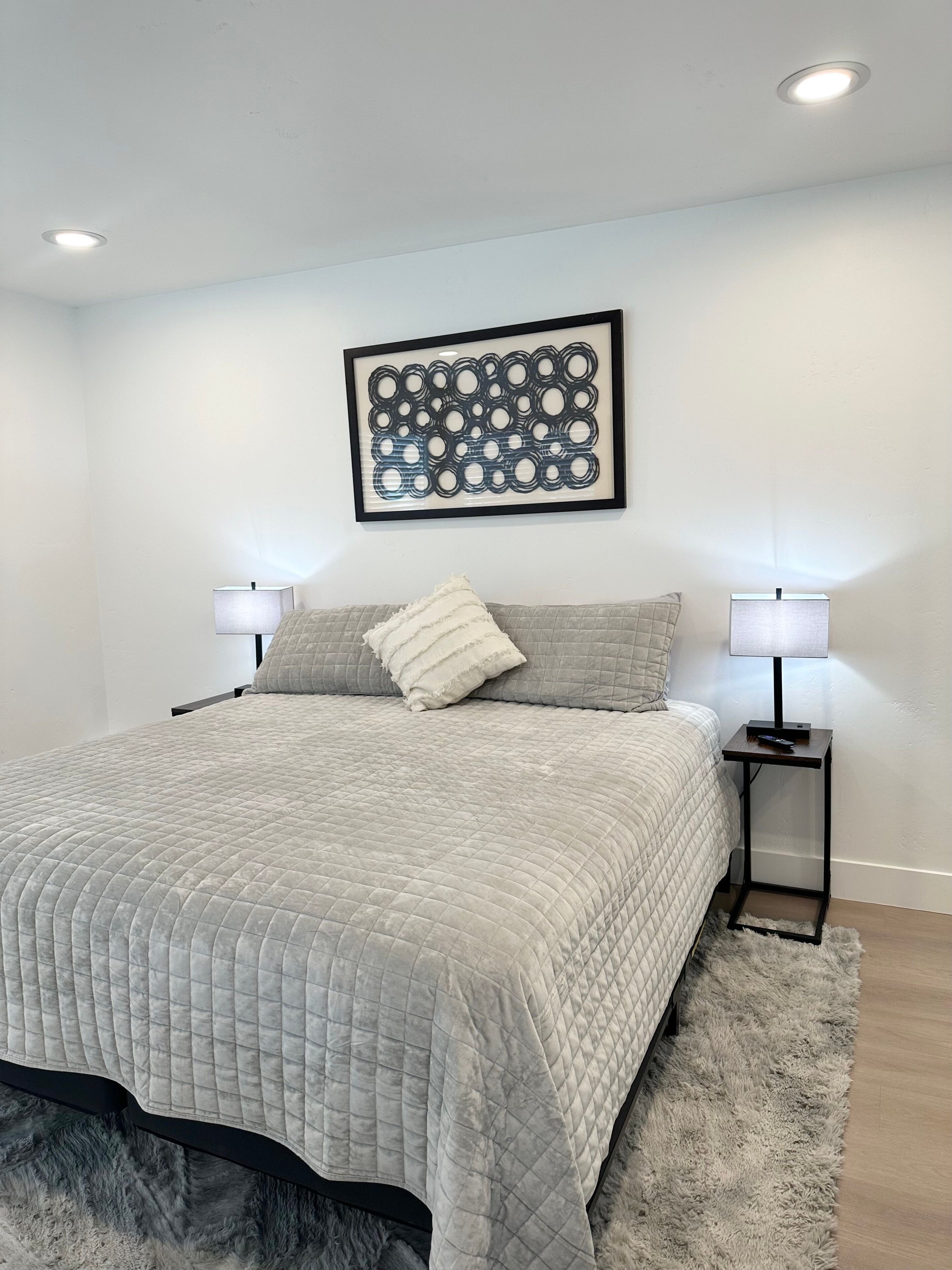 A bedroom with a bed, two bedside tables with lamps, a framed abstract artwork, and a fluffy rug.