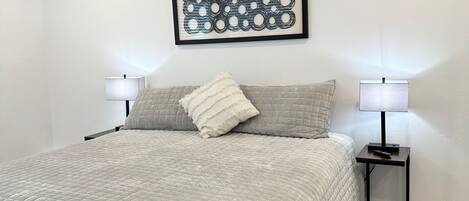 A bedroom with a bed, two bedside tables with lamps, a framed abstract artwork, and a fluffy rug.