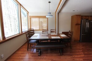 A dining area with a wooden table, chairs, and a bench. There is a window with blinds, a hanging light fixture, and a refrigerator in the kitchen area. - Coh1261 - Nature's Gem 4 Bedroom Home (South Lake Tahoe)