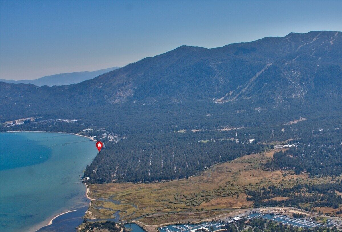 An aerial view of a coastal area with a marina, forested hills, and a clear sky.