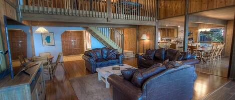 A spacious living area with a high ceiling, wooden flooring, a staircase leading to an upper level, and a dining area visible through a glass door.
