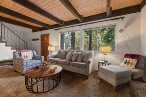 A cozy living room with a sofa, armchair, ottoman, and coffee table. A staircase with a wooden railing is visible in the background. - Coh0885 - Skyline Divine 4 Bedroom Home (South Lake Tahoe)