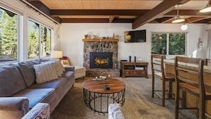 A cozy living room with a stone fireplace, a sofa, a coffee table, and a dining area with wooden chairs and a table. - Coh0885 - Skyline Divine 4 Bedroom Home (South Lake Tahoe)