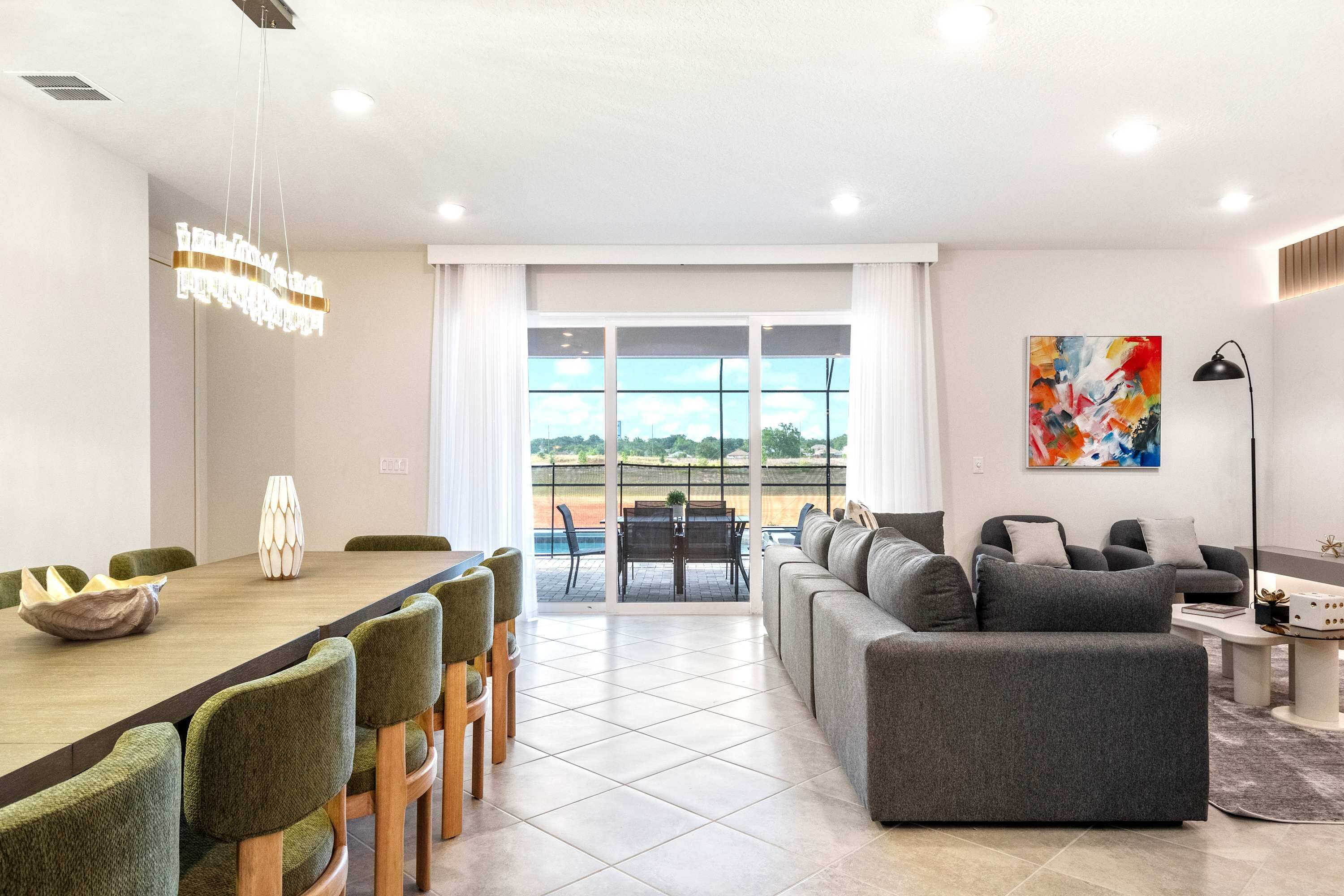 A modern living room with a dining area, a gray sofa, and a view of a tennis court through large windows.