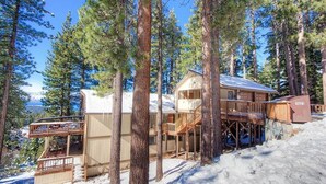 Una cabaña de madera con balcón y terraza, rodeada de altos pinos y nieve.