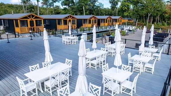 A deck with white tables and chairs, a pool, and wooden cabins in the background.