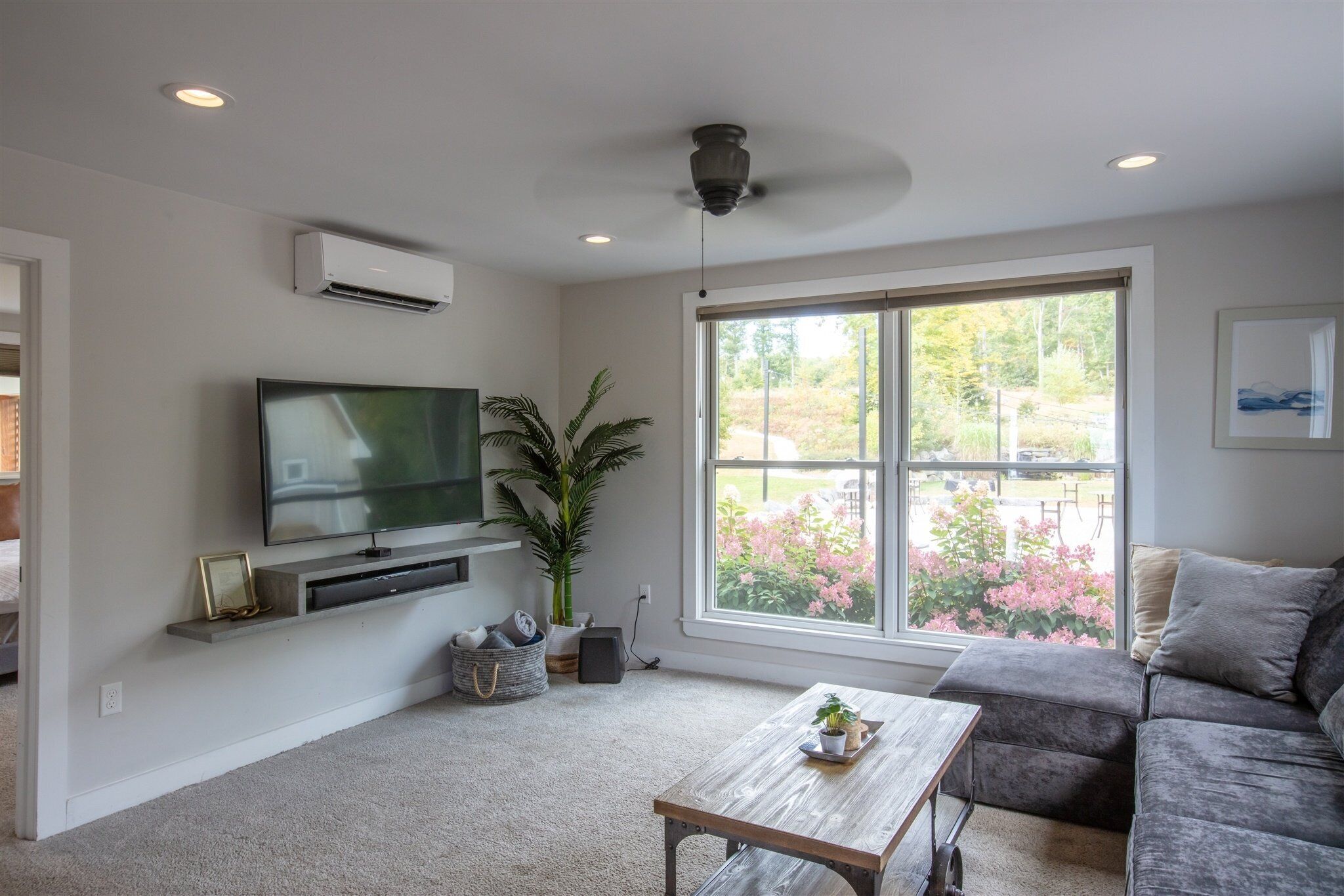A living room with a flat-screen TV on a wall-mounted shelf, a large window with a view of greenery, a grey sofa, and a wooden coffee table.