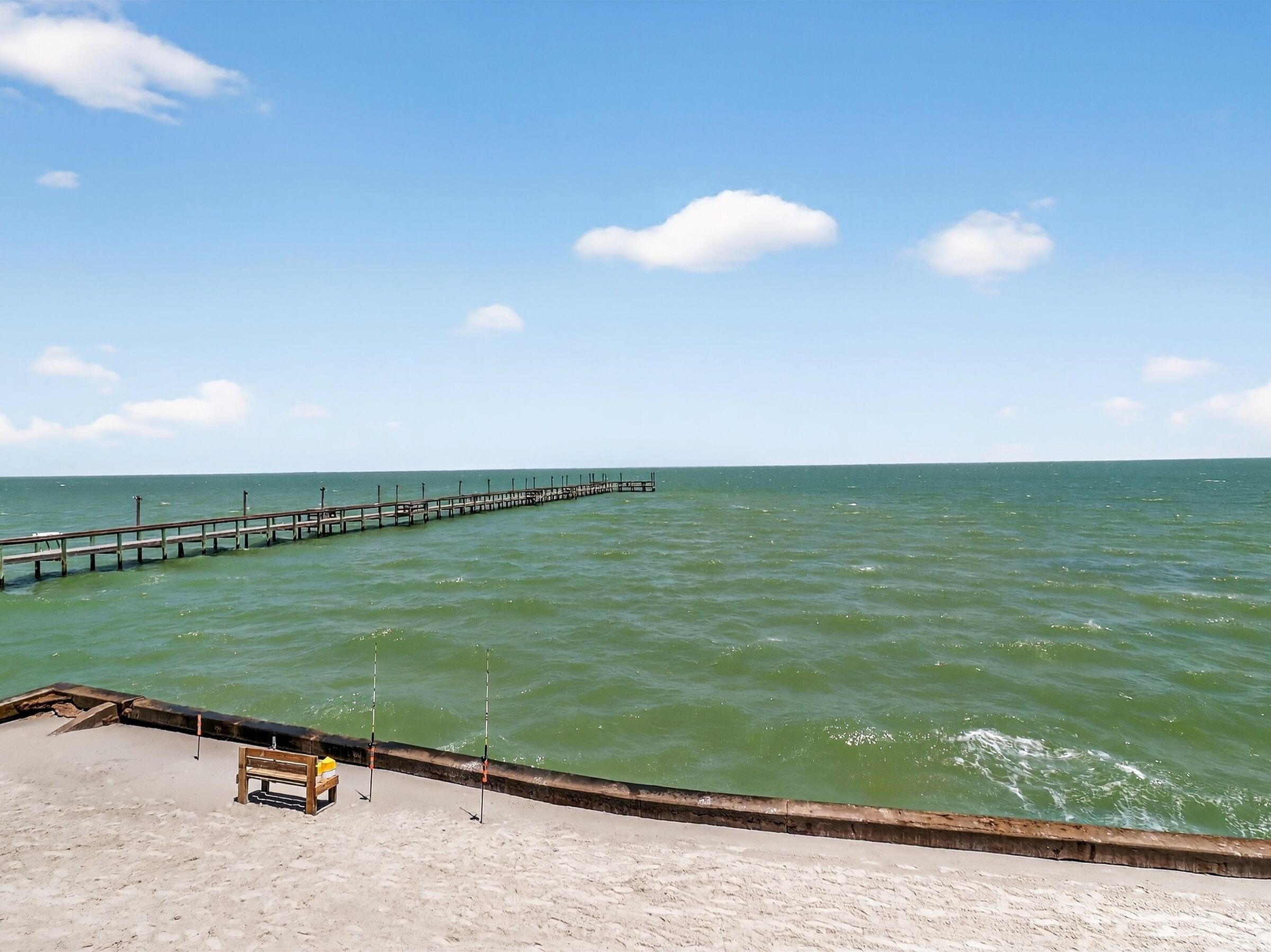 A pier extending into calm waters with a sandy beach and a bench in the foreground.