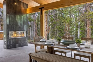 A modern outdoor dining area with a built-in fire feature, wooden furniture, and a view of a forested area. - Saddle Horn Lane 4 Bedroom Home (Telluride)