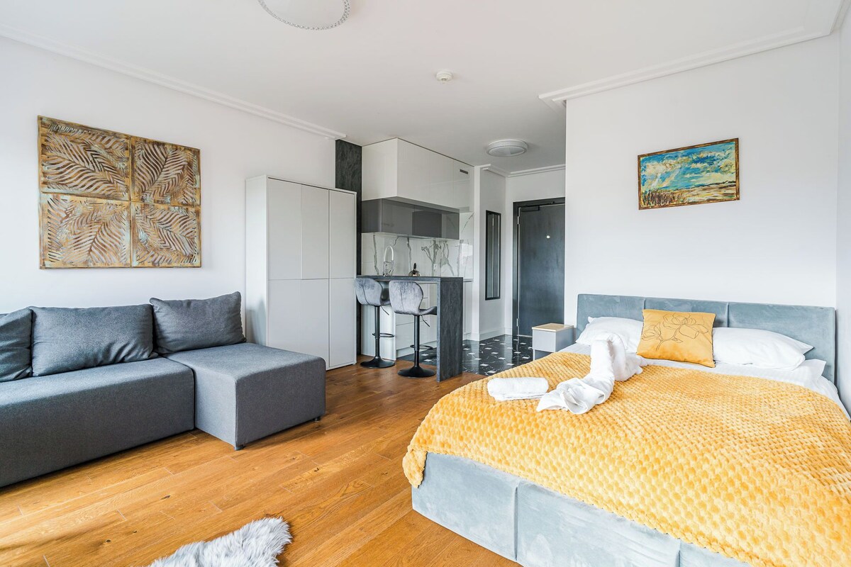 Une chambre à coucher moderne avec un canapé gris, un lit recouvert d’une couverture jaune et, au fond, un coin cuisine.