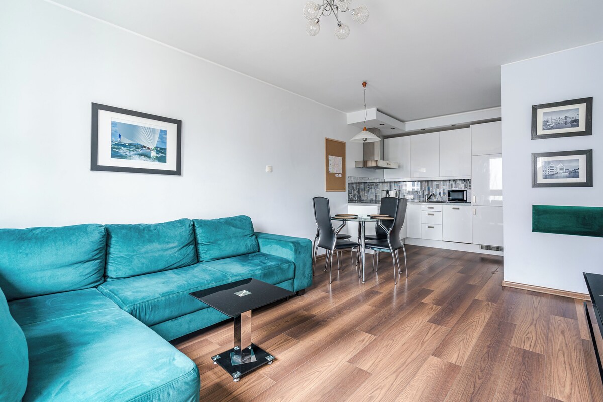 Un salon moderne avec un canapé de couleur turquoise, une petite table noire et un coin repas avec des chaises. On aperçoit une cuisine à l’arrière-plan, avec des meubles blancs et une hotte de cuisinière en acier inoxydable.