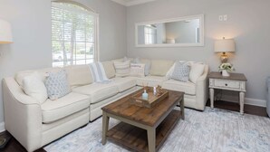 Ein Wohnzimmer mit einem Ecksofa, einem Holztisch, einem Beistelltisch mit Lampe, einem Deckenventilator und einem großen Fenster mit Jalousien.