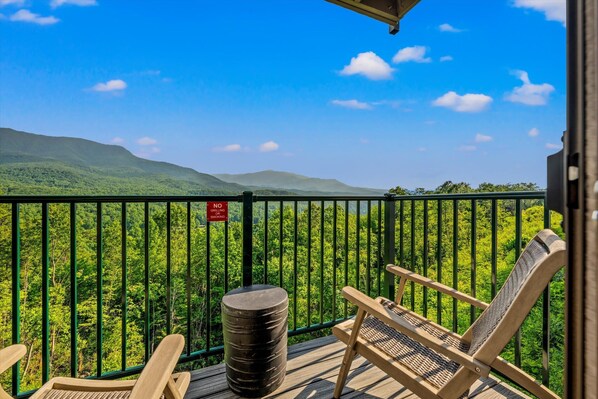 A balcony with a view of a forested mountain range, a no grilling sign, and two wooden chairs.