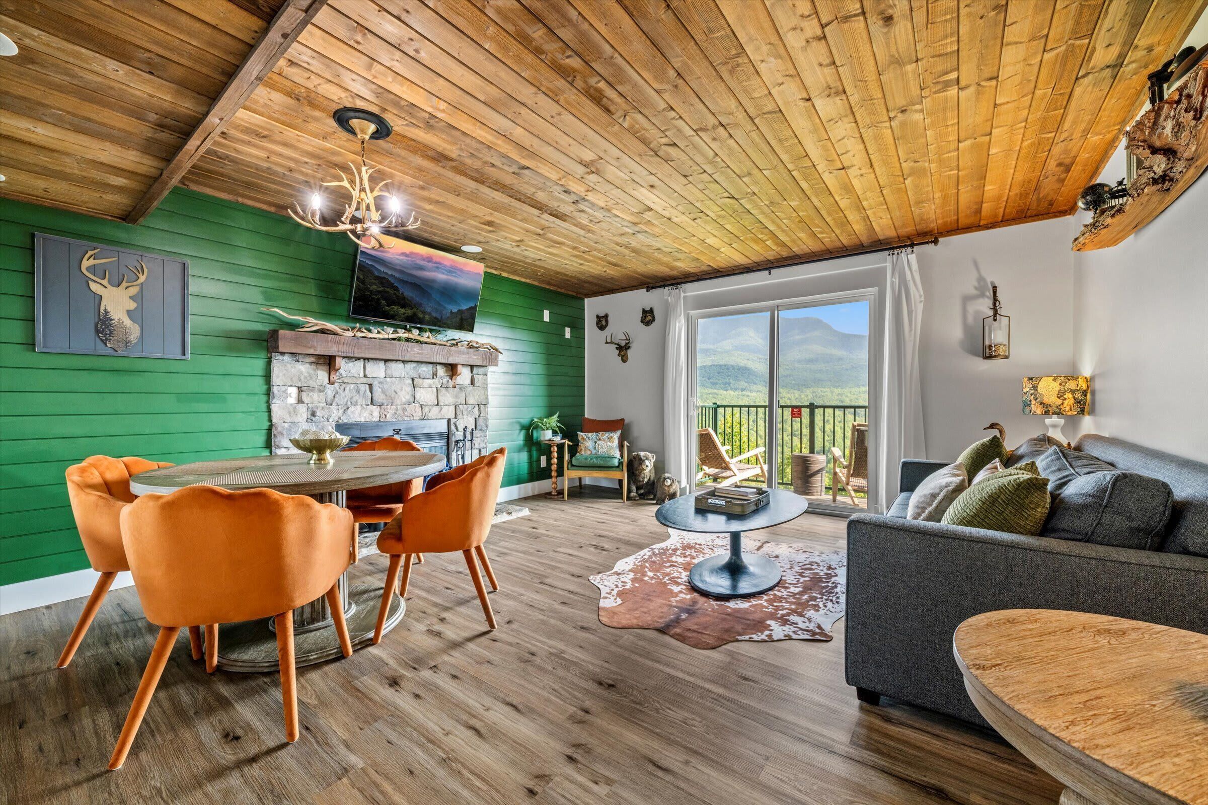 Ein gemütliches Wohnzimmer mit Holzbalkendecke, grünen Wänden, einem Kamin aus Stein und einem Essbereich mit orangefarbenen Stühlen.