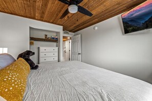A bedroom with a bed, a ceiling fan, a white dresser, a shelf with books, and a door leading to another room.
