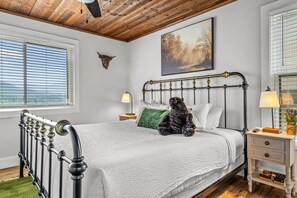 Un dormitorio con estructura de cama metálica, ropa de cama blanca, almohada verde, mesita de noche de madera con una lámpara y un cuadro en la pared.