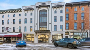 A multi-story building with a central entrance, storefronts on the ground floor, and a parking lot with cars. - Local Discounts!-The Revitalized Newark Arcade (Newark)