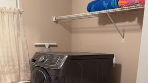 A laundry room with a black and a white front-loading washer and dryer, a window with curtains, and a shelf above the dryer holding folded clothes. - “Welcome to this charming 3bedroom home in a quiet cul-de-sac in Evans Ga. (Evans)