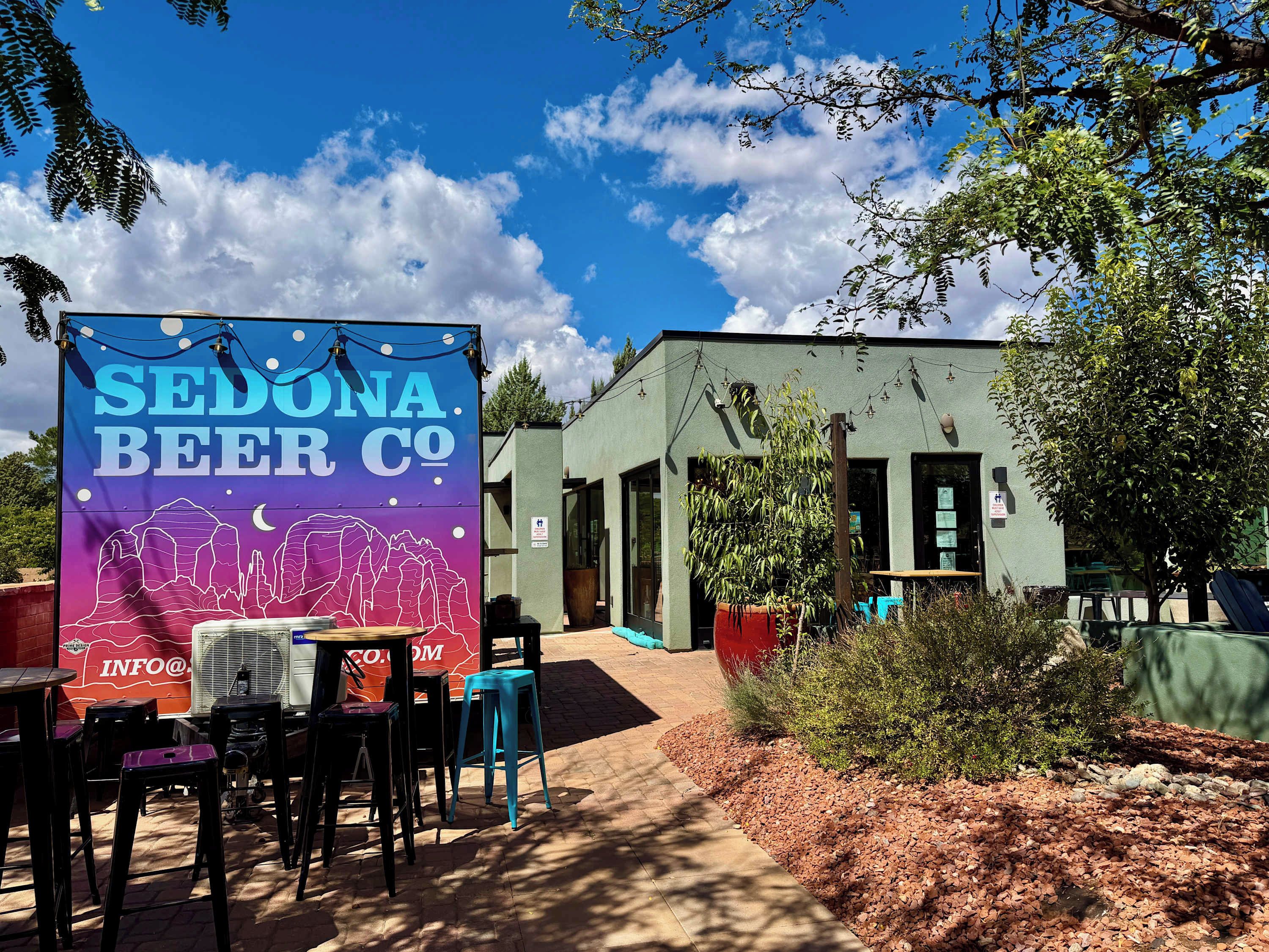 Ett utomhusområde med bord och stolar, en banderoll för Sedona Beer Co. och en byggnad med en grön fasad.
